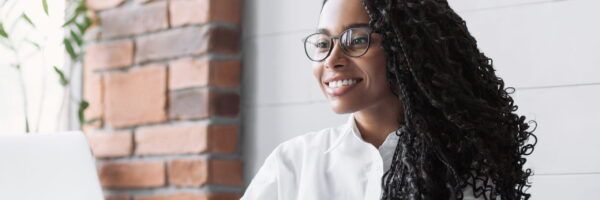 A smiling young woman with glasses and long curly hair specializing in medical billing and coding, sitting at a desk with a laptop, looking off to the side in a modern office setting.
