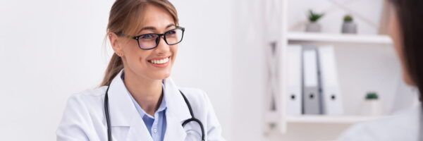 A smiling female doctor with glasses, wearing a white lab coat and a stethoscope, converses with a patient in a bright, modern office about budgeting for school expenses.