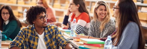 A diverse group of university students engaging in discussion in a classroom, with books and water bottles on the desks. They appear cheerful and attentive as they analyze common freshman mistakes.