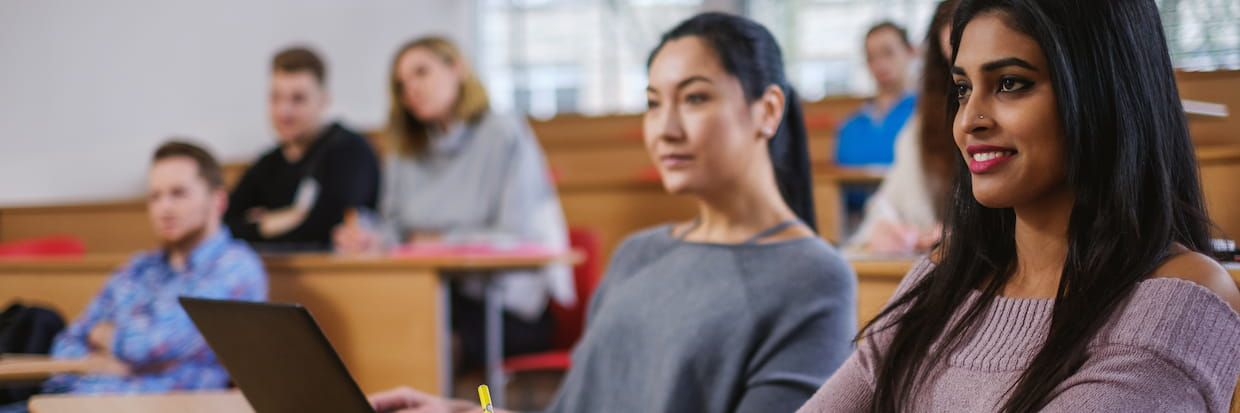 Diverse group of college students sitting in a lecture hall, focusing intently ahead. A woman in the foreground smiles slightly while using a laptop.