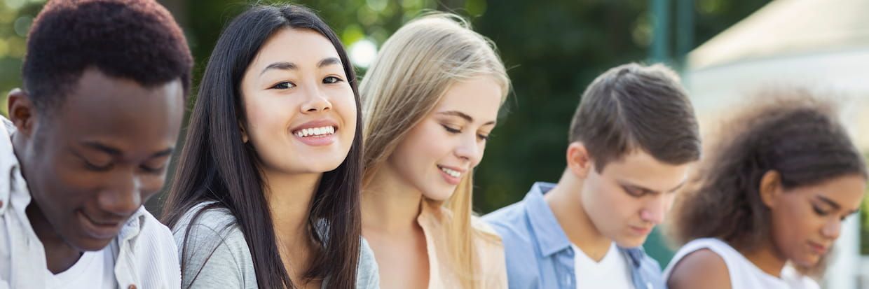 A group of diverse college students standing outdoors, including an Asian woman smiling at the camera, with others focused on their phones.