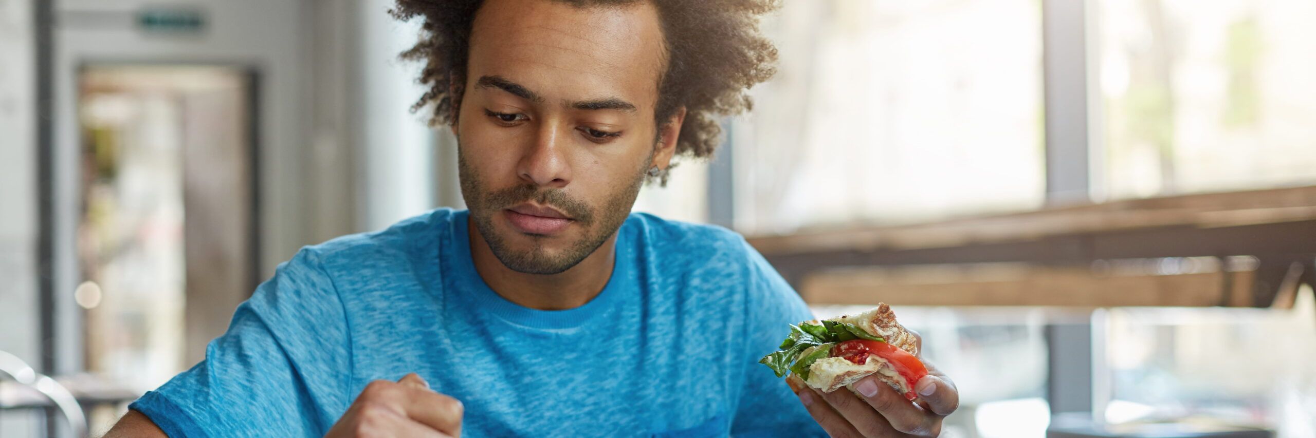 A college student in a blue shirt attentively examines a sandwich while sitting in a brightly lit cafe.