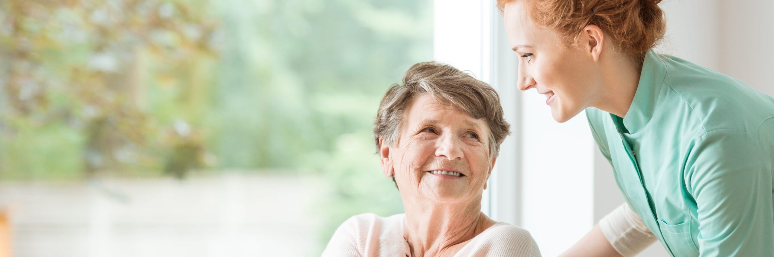 A caregiver, a young woman with red hair, gently smiles at an elderly lady who is sitting and looking back at her with a joyful expression, both near a bright window with greenery outside. They are navigating healthcare challenges together.