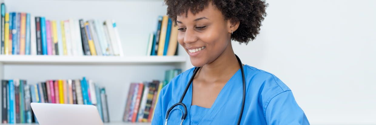 A cheerful female healthcare professional in blue scrubs using a tablet in front of a bookshelf filled with colorful books on medical school admission.