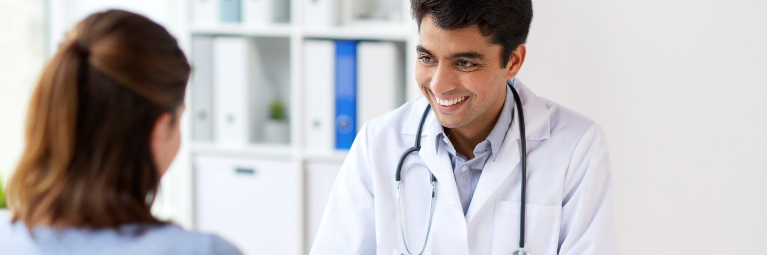 A smiling male doctor in a white coat with a stethoscope around his neck is engaging with a female college student in a medical office.