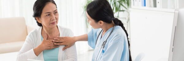 A healthcare professional attentively listens to an elderly woman who is touching her throat, indicative of explaining a symptom related to medical coding, in a clinical setting.