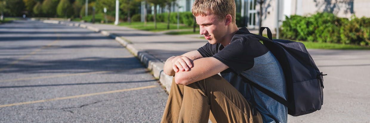A college student with short blonde hair, wearing a gray shirt and khaki pants, sits on a curb in an empty parking lot. He looks pensively to the side while carrying a black backpack.