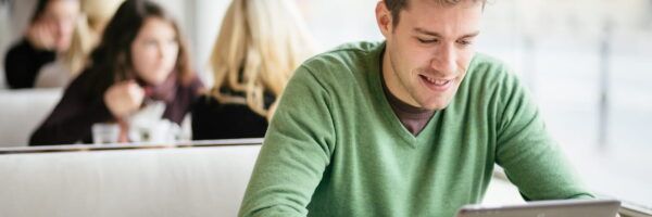 A young man in a green sweater smiling while focusing on a tablet, seated in a brightly lit cafe studying for the MCAT with other people in the background.