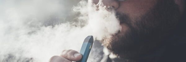 Close-up of a bearded man exhaling vapor from an electronic cigarette, focusing on the device in his hand and the cloud of vapor surrounding him as he attempts to quit smoking.