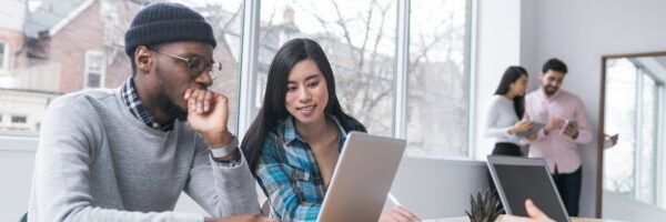 Two young professionals, a man in a beanie and glasses and a woman with long hair, collaborating on transferring an allied health license using a laptop in a modern office, with two colleagues discussing in the