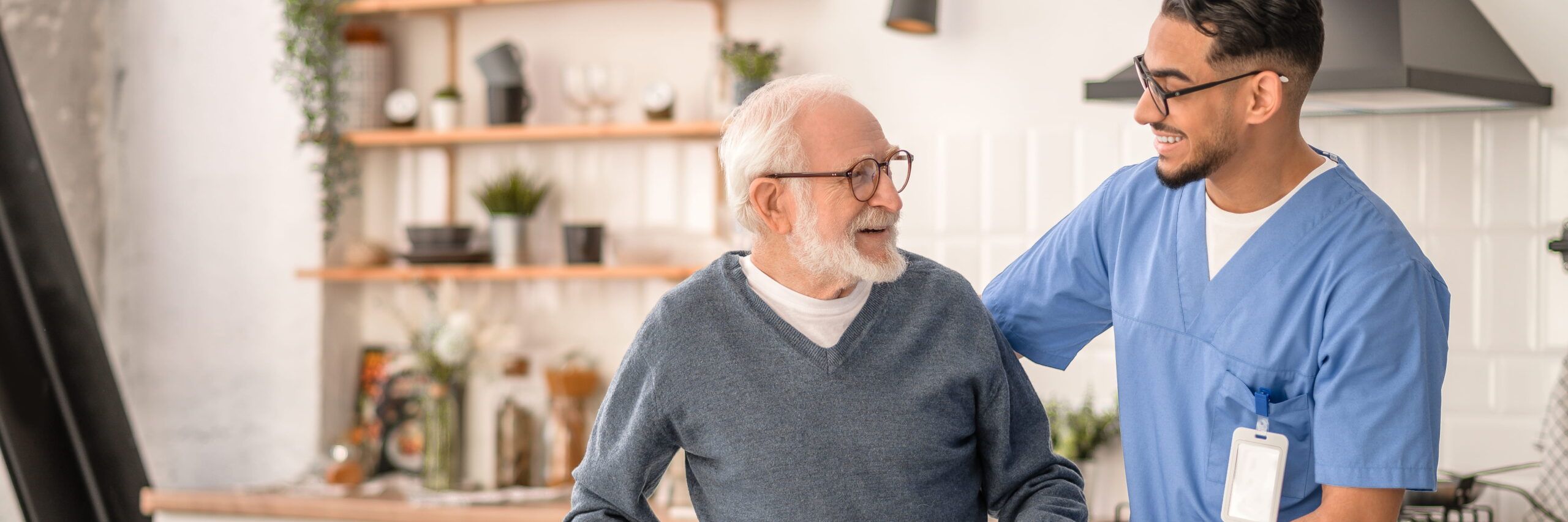 A senior man with glasses and a young male nurse in scrubs, both smiling as the male nurse assists the man in a warmly lit room filled with plants.