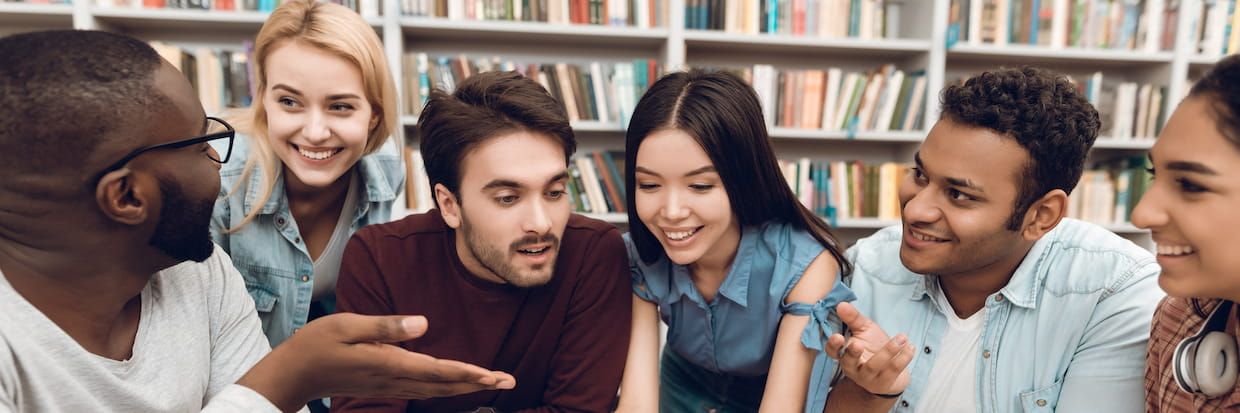 A diverse group of six young adults, including minority students, engaged in a lively discussion about healthcare education in a library, with bookshelves filled with books in the background.