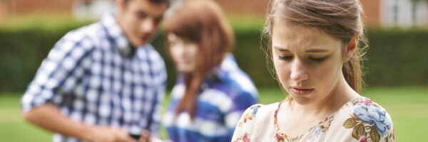 A young woman looking distressed in the foreground while a teenage boy and girl talk behind her, possibly excluding her, in an outdoor setting related to a Master's in Nursing program.