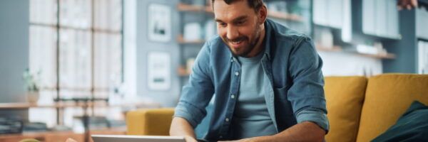 A joyful man with a beard working on a laptop while sitting on a yellow sofa in a cozy, modern living room, researching SEO keywords.