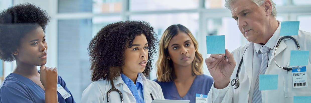 Three healthcare professionals, two women and an older man, discussing summer research opportunities over notes in a bright hospital corridor.