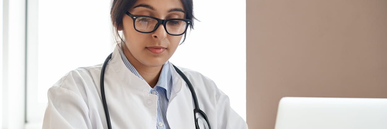 A female doctor in glasses wearing a white coat with a stethoscope around her neck, concentrating on her laptop screen in a bright office setting.