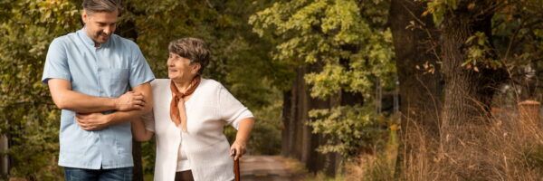 A smiling male medical volunteer in blue scrubs assists an elderly woman as they walk together in a park, surrounded by autumn trees.