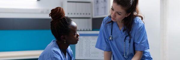 Two healthcare professionals, one sitting and one standing, engaged in a serious discussion over a nursing assignment in a clinical setting.