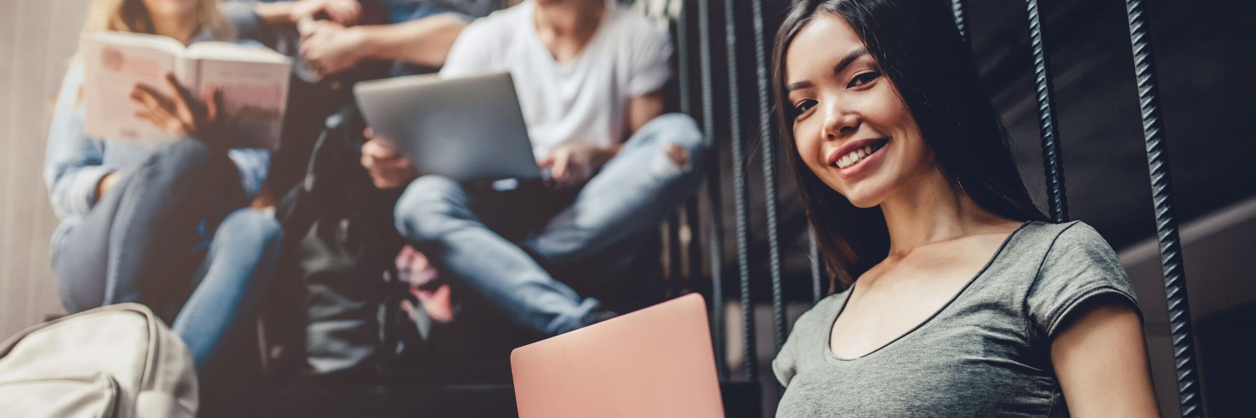 A smiling young asian woman holding a pink folder, sitting near a group of students engaged in reading and using a tablet, indoors with a modern, casual vibe.