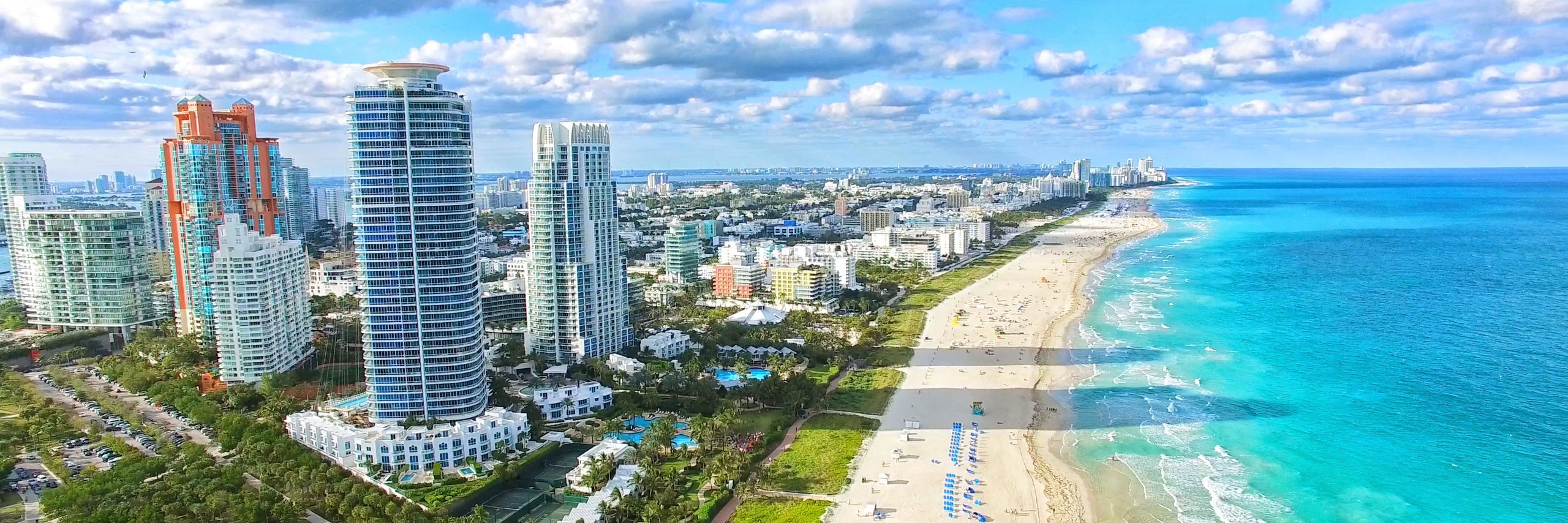 Aerial view of a vibrant coastal cityscape with tall skyscrapers adjacent to a sandy beach along a clear turquoise ocean under a partly cloudy sky, featuring renowned massage therapy centers.