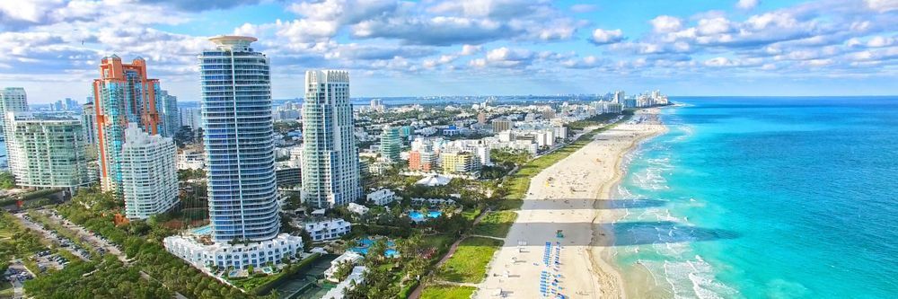 Aerial view of a coastal city with skyscrapers beside a sandy beach, extending toward a clear blue ocean on a sunny day, with a lush green park visible in the foreground.