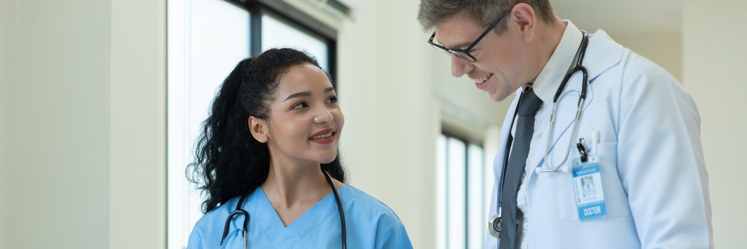 Two healthcare professionals, one man and one woman, are conversing and smiling in a hospital corridor. both are wearing scrubs and stethoscopes.