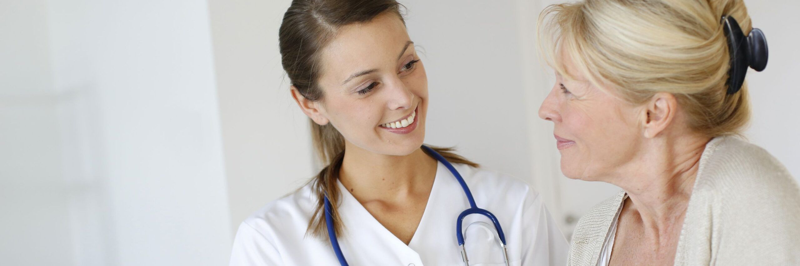 A female healthcare professional with a stethoscope smiling at an older female patient wearing a sweater, both seated in a medical office.