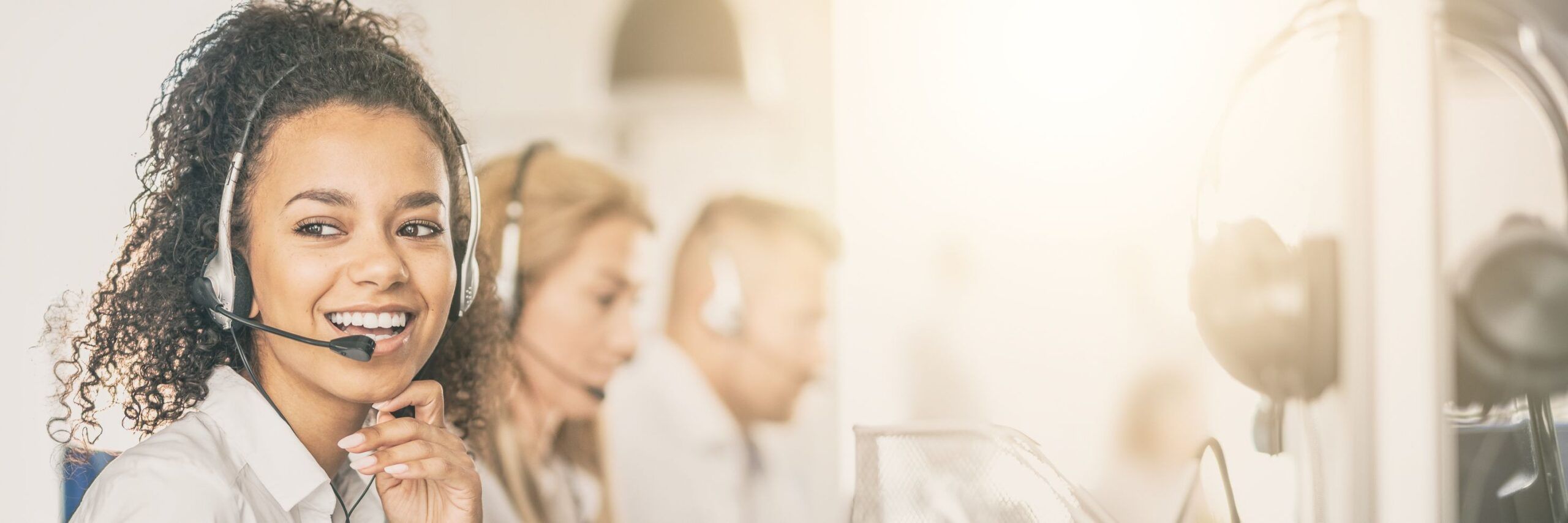 Smiling woman with curly hair wearing a headset in a bright call center, colleagues in background also wearing headsets and working.