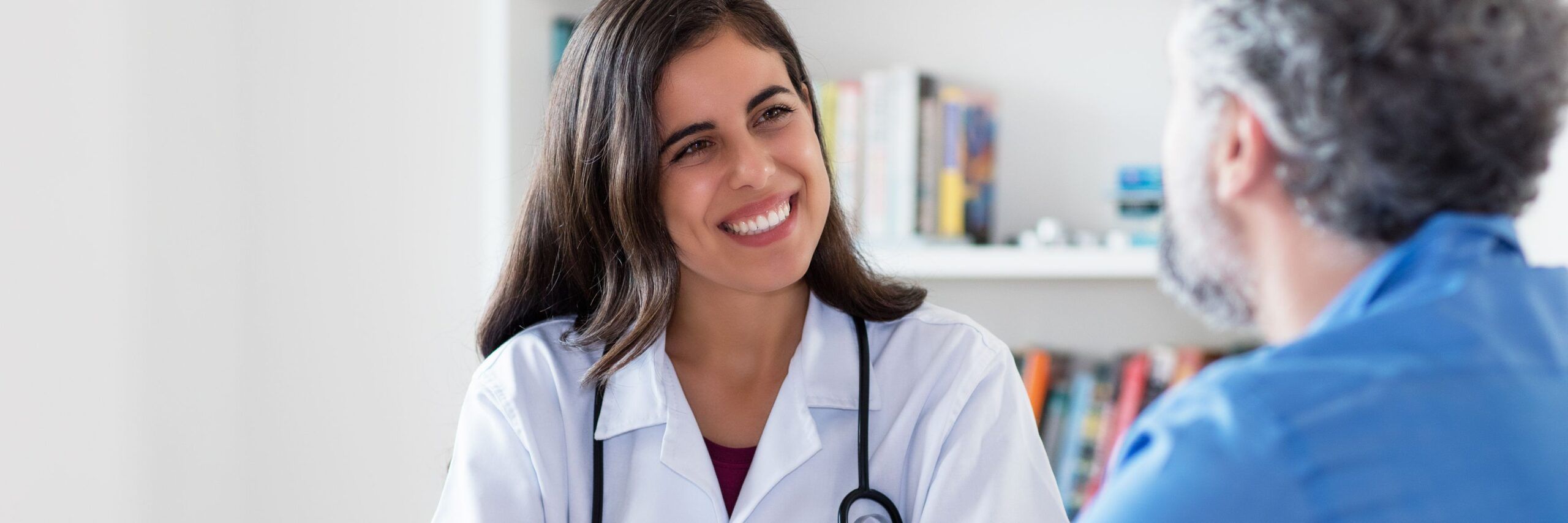 A female doctor with dark hair, smiling, talking to a male patient with gray hair in a medical office. she wears a white coat and a stethoscope around her neck.