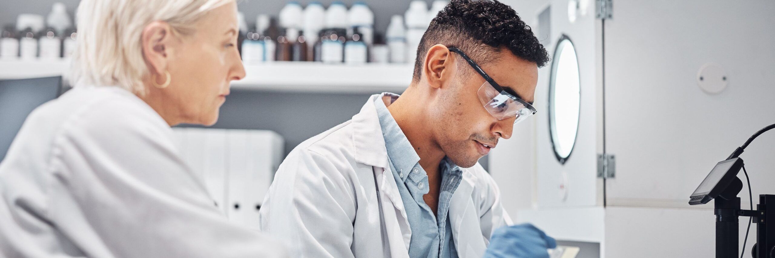 Two scientists, a young man and an older woman, wearing lab coats and eyeglasses, engaged in research work in a laboratory, examining samples with a microscope.