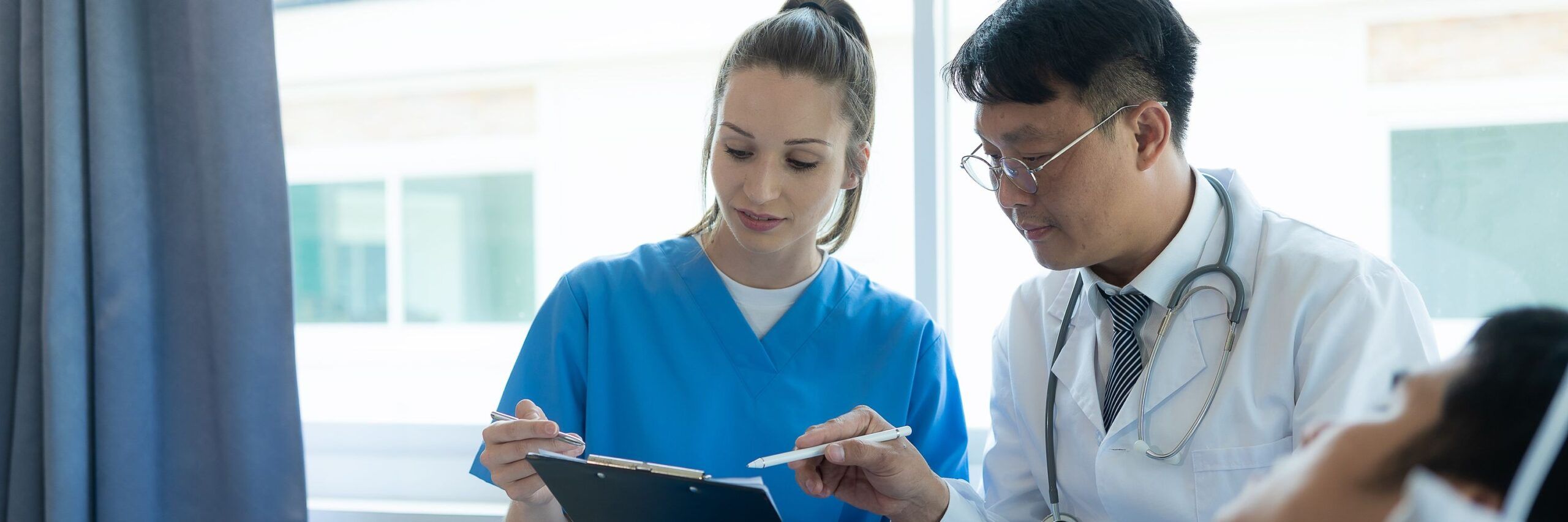 Two healthcare professionals, a man in a white coat and a woman in blue scrubs, are discussing a document on a clipboard in a hospital setting.