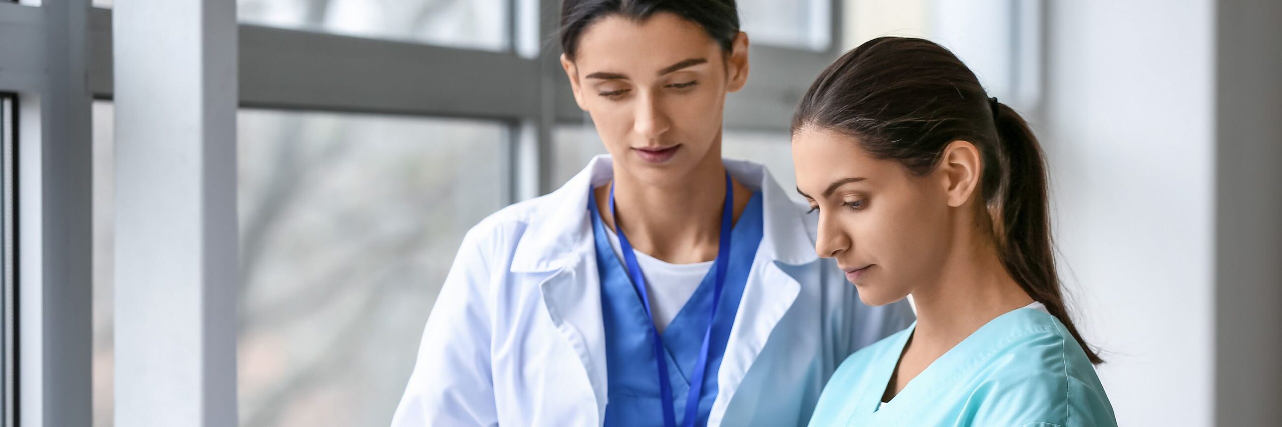 Two female healthcare professionals, one wearing a white coat and another in blue scrubs, are walking and discussing in a hospital corridor with large windows.