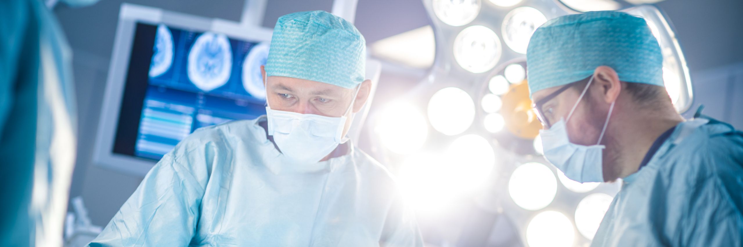 Two surgeons in scrubs and surgical masks focus intently during a procedure in an operating room illuminated by bright surgical lights, with medical monitors in the background.