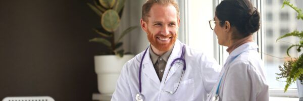 A smiling male doctor with a beard, in a white coat and stethoscope, converses with a female colleague in scrubs in a brightly lit office.