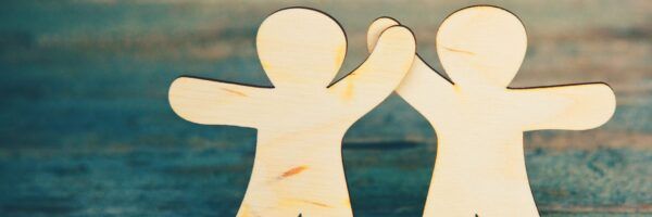 Two wooden cutout figures resembling people stand side by side against a blurred blue and brown background, symbolizing friendship or partnership.