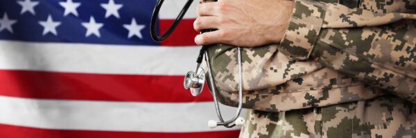 A close-up of a person in military camouflage uniform holding a stethoscope, with the american flag in the background.
