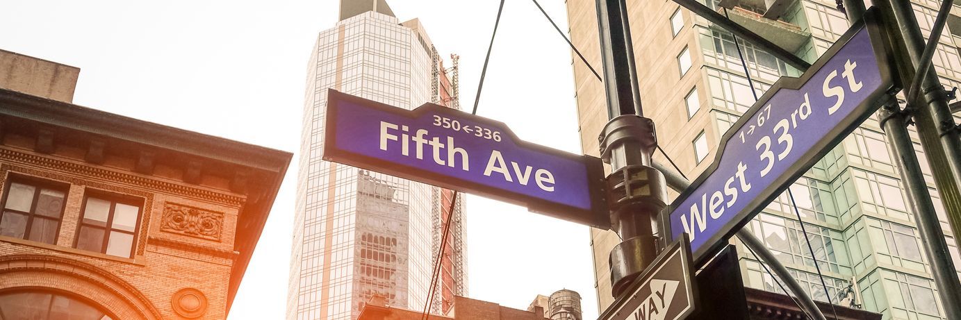 Street signs for Fifth Avenue and West 33rd Street in Manhattan, New York, with a view of high-rise buildings and a construction crane under a bright, hazy sky. Visit Georgia.