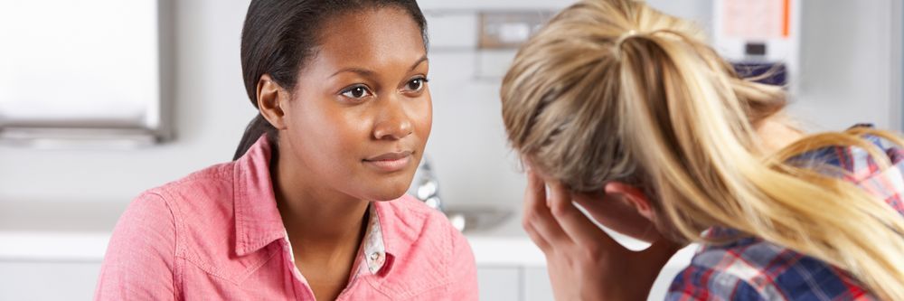 Two women sitting at a kitchen counter engaged in a serious conversation, with one looking concerned and the other offering a comforting gaze.