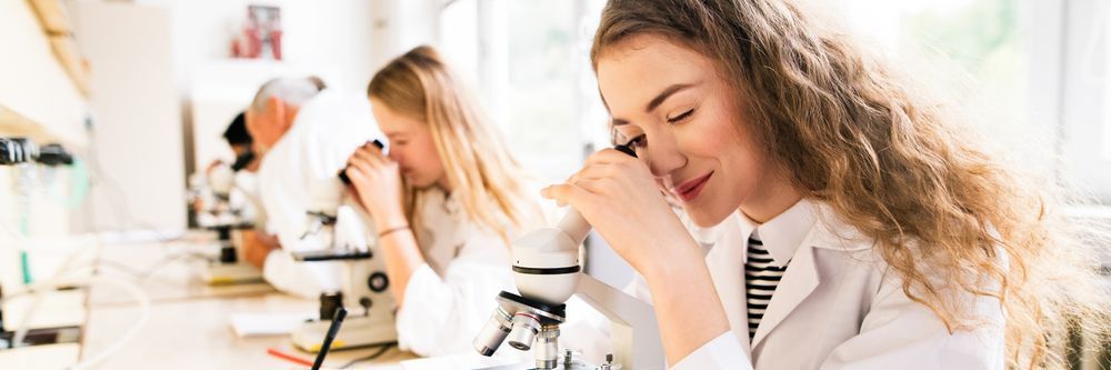 Young female scientist using a microscope in a laboratory with other researchers working in the background.