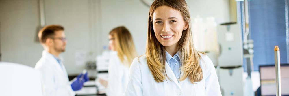 A smiling young woman in a lab coat stands in a laboratory. in the background, two other scientists are engaged in their work. the setting is bright and modern.