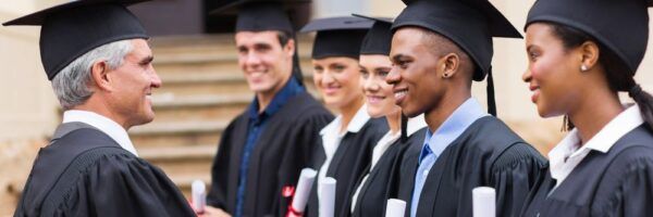 A professor congratulating a line of diverse graduates in cap and gown, each holding a diploma, outside a university building.