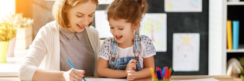 A smiling woman helps a young girl with drawing at a brightly lit table filled with colorful art supplies in a cheerful educational setting.