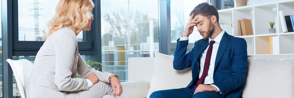 A man in a blue suit sits with his head in his hand, looking distressed, while a woman opposite him listens attentively in a bright, modern office setting.