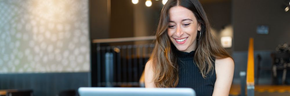 A smiling woman using a laptop at a cafe with a stylish, modern interior. she is focused on the screen, wearing a black sleeveless top.