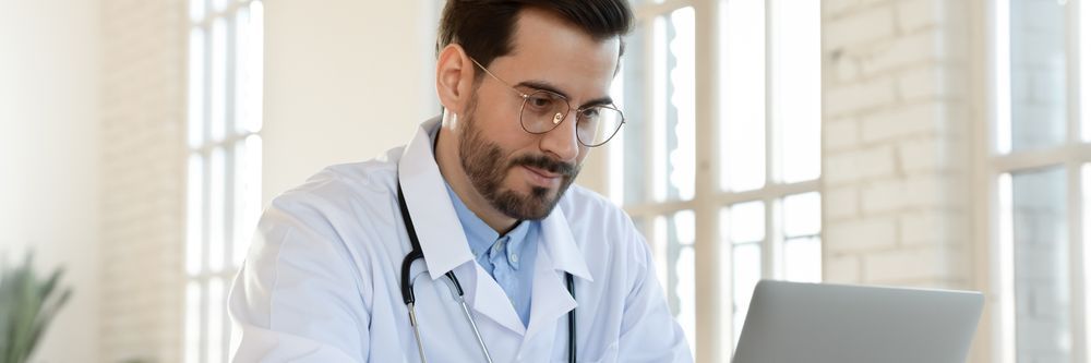 Male doctor with glasses and a stethoscope, wearing a white coat, looks at a laptop, seated at a desk in a bright, sunlit office.