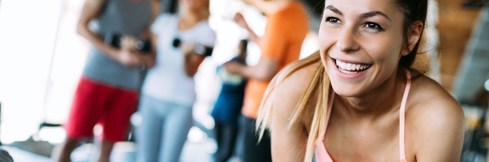 A joyful woman in workout gear smiling at the camera with a group of people exercising in the background at a gym.