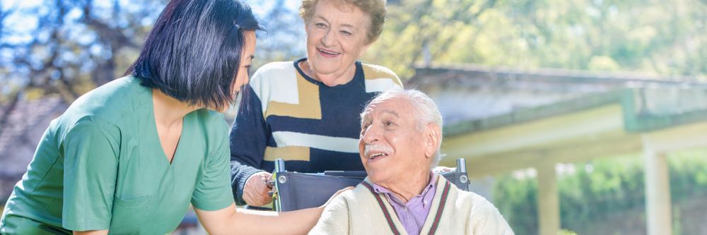 A female nurse outdoors assisting a smiling elderly man in a wheelchair, with an elderly woman standing alongside, all engaging in a cheerful conversation.