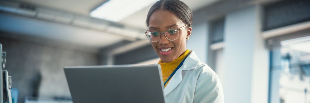 A smiling black woman in a lab coat and glasses using a laptop in a modern office setting, suggesting a professional or scientific environment.