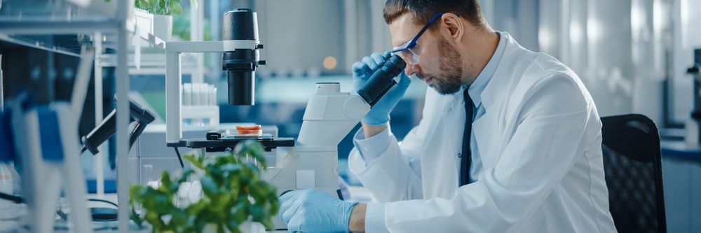 A male scientist in a lab coat and gloves examines a sample using a microscope in a well-equipped laboratory.
