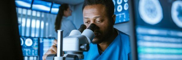A focused male scientist in a blue lab coat examines samples through a microscope in a high-tech lab, with a female colleague working in the background.